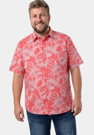 Homme souriant avec barbe portant une chemise corail rose à manches courtes avec un motif léger de feuilles et un jean bleu, debout devant un fond clair uni.
