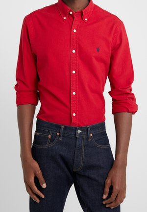 Homme portant une chemise rouge vif à boutons avec les manches retroussées et un jean bleu foncé, debout devant un fond clair uni.