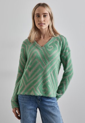 Kvinna som bär en grön och beige mönstrad mjuk tröja med V-ringning och blå jeans mot en enkel ljusgrå bakgrund.