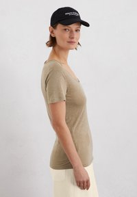 Beige, aansluitend t-shirt met een v-hals en korte mouwen, gecombineerd met een zwarte pet met witte tekst. Soepele stofstructuur.