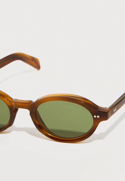 Lunettes de soleil ovales à verres verts avec une monture translucide en écailles de tortue marron et deux petites clous métalliques en forme d'étoile près des charnières.