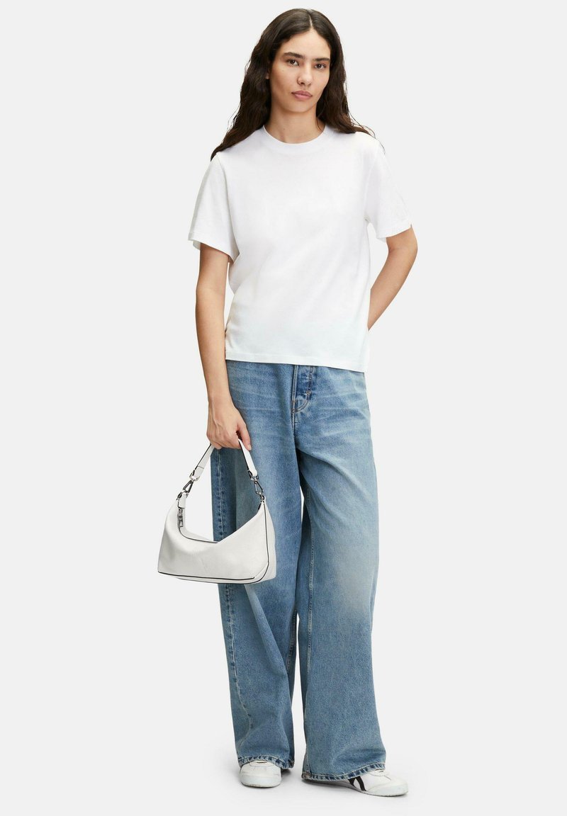 T-shirt blanc à manches courtes associé à un pantalon en jean bleu clair à jambes larges. Un petit sac à main blanc est tenu dans une main. Des baskets blanches complètent le look.