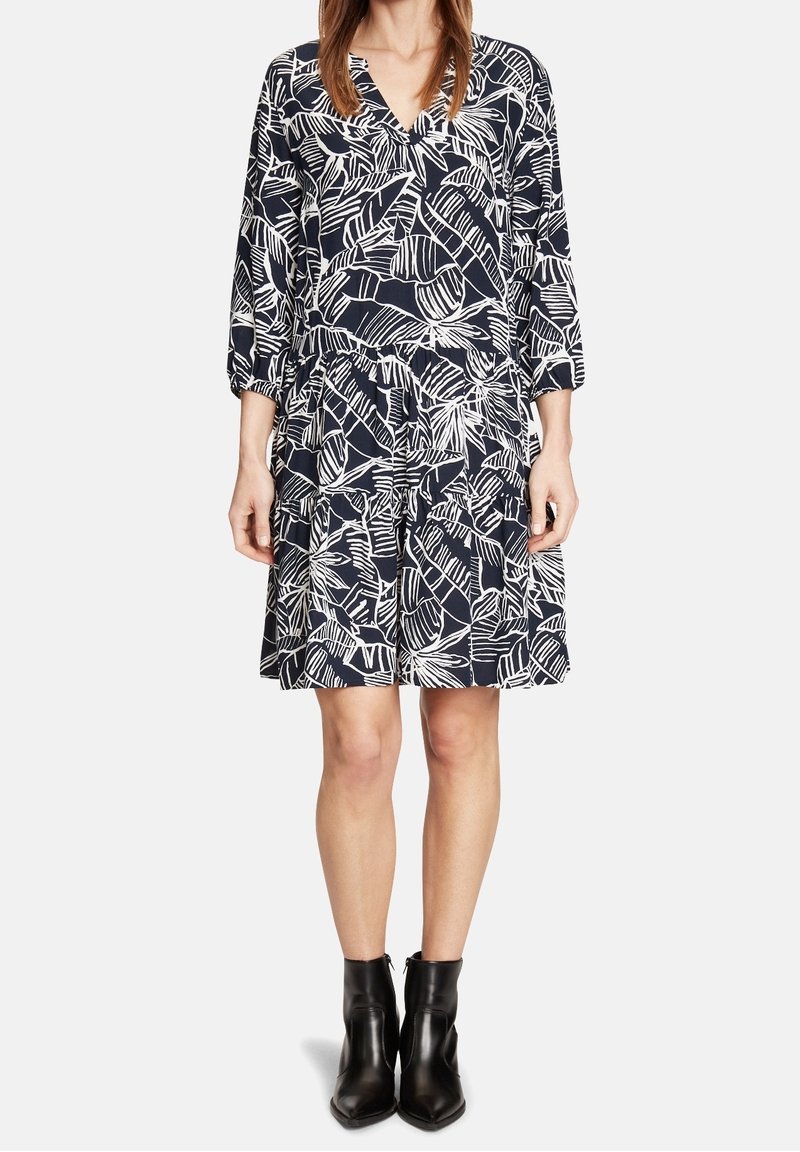 Robe marine avec impression de feuilles blanches, décolleté en V et manches trois-quarts. Dispose d'une jupe froncée et d'un tissu texturé. Portée avec des bottines noires.