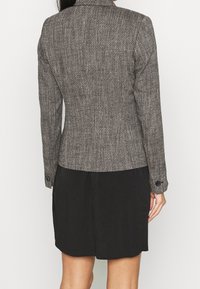 Rakenteinen, kudottu bleiseri harmaan sävyissä, jossa on istuva leikkaus, lovikaulus ja mustat nappikoristeet, yhdistetty yksiväriseen mustaan mekkoon.