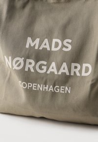 Μπεζ υφασμάτινη τσάντα με λευκή εκτύπωση που γράφει "MADS NØRGAARD COPENHAGEN" πάνω σε λευκή επιφάνεια.