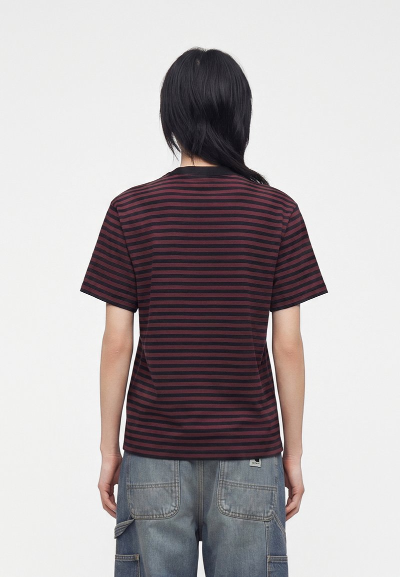Persona con capelli lunghi e neri che indossa una maglietta nera a righe rosso scuro e jeans blu slavati larghi, vista da dietro.