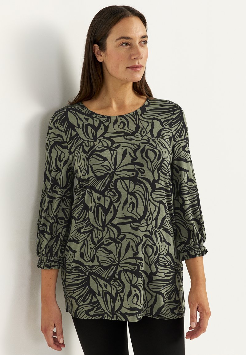 Frau mit braunen Haaren, die eine grüne Bluse mit schwarzen abstrakten Blumenmustern und Dreiviertelärmeln trägt, steht vor einer weißen Wand.