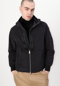 Black hooded jacket with a textured finish, front zipper, and two large chest pockets featuring black accents. Paired with a black turtleneck.