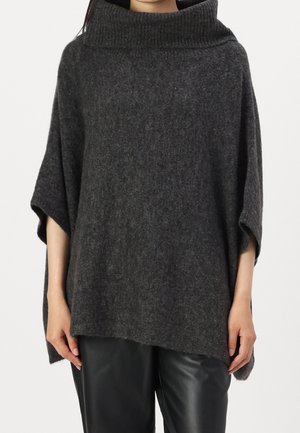 Personne portant un poncho ample en laine gris foncé avec un large col roulé côtelé et un pantalon en cuir noir, debout, les bras détendus.