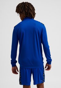 Blaues sportliches Langarm-Top mit strukturierter Stoffoberfläche und gerippten Bündchen, kombiniert mit passenden Shorts mit einem gemusterten Seitenstreifen.