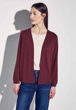 Burgunderfarbener Cardigan mit lockerer Passform, offener Vorderseite und langen Ärmeln. Kombiniert mit einem cremefarbenen Oberteil und blauen Jeans, die sichtbare Denim-Details aufweisen.