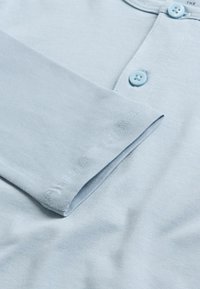 Svijetloplava pamučna majica s henley dizajnom, dva usklađena gumba na izrezu vrata i glatkom teksturom. Rukavi su blago prevrnuti.