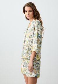 Ensemble de détente floral comprenant un haut à manches courtes avec des boutons et un short assorti. Tissu léger avec des motifs botaniques verts, jaunes et gris.