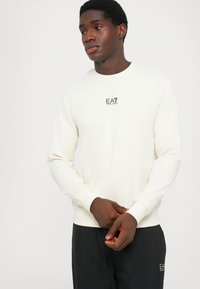 Sudadera de color beige claro hecha de tejido suave, con cuello redondo, mangas largas y un pequeño logo negro en el pecho.