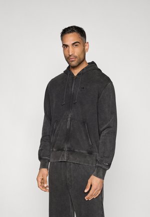 Homme portant un sweat à capuche zippé gris foncé avec cordons de serrage et un pantalon de survêtement assorti, debout devant un fond clair uni.