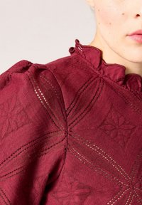 Burgundin värinen teksturoitu paita, jossa on röyhelökaulus ja monimutkaisia geometrisia kuvioita leikkuuleikkaus yksityiskohdilla. Pehmeä kangas, jossa on ylellinen pinta.