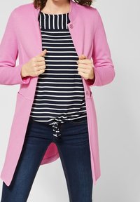 Vaaleanpunainen pitkä neuletakki, jossa on pyöreä pääntie ja yksi nappikiinnitys, käytetään musta-valkoisen raidallisen topin päällä. Yhdistetty tummiin farkkuihin.