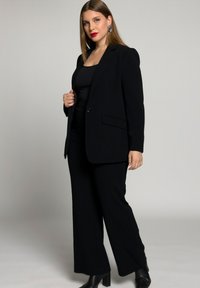 Completo nero su misura con un blazer strutturato e pantaloni a gamba larga, abbinato a un top aderente, dalla texture lucida, e stivali neri con tacco spesso.