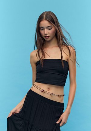 Top bandeau nero corto abbinato a una gonna plissettata nera. Una catena decorativa di perline multicolori avvolge la vita. Lo sfondo è azzurro chiaro.