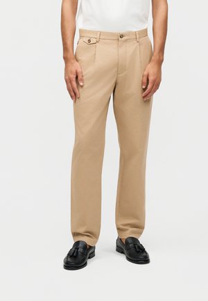Mand iført beige plisserede bukser med knapdetalje på sidelomme og sorte tassel loafers, stående mod en hvid baggrund.