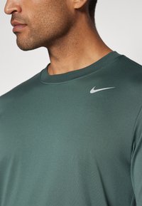 Tmavě zelené sportovní tričko s kulatým výstřihem, měkkou texturou a malým bílým logem Nike swoosh na levém rameni.