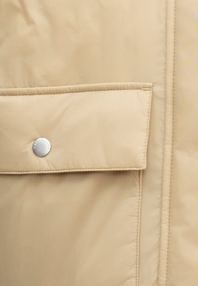 Beežikas pufferjope, millel on klapiga tasku ja hõbedane kinnitusnupp. Sile viimistlus nähtavate õmblustega ja minimalistlik disain.