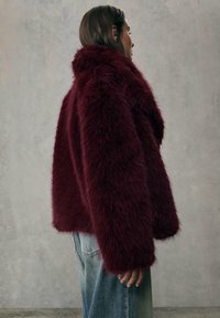 Burgundowa kurtka z sztucznego futra z wysokim kołnierzem, luźnym krojem i surowym wykończeniem, połączona z jasnoniebieskimi dżinsami, prezentująca miękką, teksturowaną powierzchnię.