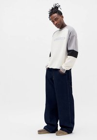 Felpa grigio chiaro, nera e bianca con la scritta "TRANS"; abbinata a jeans larghi in denim scuro; calzature casual completano il look.