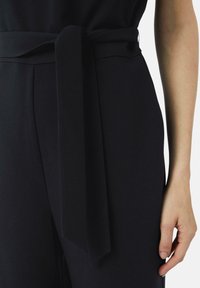 Zwarte zachte stoffen jumpsuit met een brede tailleband die aan de voorkant is gestrikt. Het materiaal lijkt soepel, met een schone, op maat gemaakte pasvorm die de silhouet benadrukt.