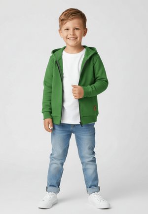Uśmiechnięty młody chłopiec stoi, ubrany w zieloną bluzę z kapturem zapinaną na zamek, białą koszulkę, jasnoniebieskie spodnie z wywiniętymi mankietami oraz białe trampki na jednolitym tle.