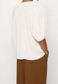 Světle růžová texturovaná blůza s nabíranými rukávy a kulatým výstřihem, nošená s hnědými kalhotami širokého střihu. Blůza má jemné záhyby.