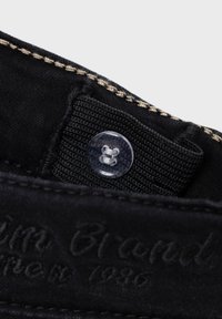 Tejido negro con costuras beige presenta un botón transparente y una etiqueta texturizada embosada con "Marca 1936". Detalle notable en la costura.