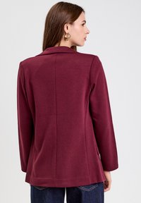 Blazer burdeos con una textura suave, solapas de muesca y dos bolsillos laterales. Presenta un corte entallado con un dobladillo ligeramente acampanado.