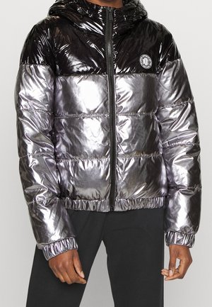 Schwarze und silberne glänzende Steppjacke mit Kapuze, Frontreißverschluss, elastischen Bündchen und einem runden Logo auf der Brust, kombiniert mit schwarzen Hosen.