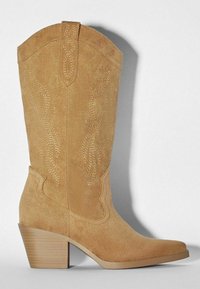 Botte de cowboy en suède beige avec un bout pointu, un talon en bois empilé et des coutures décoratives en motif tourbillonnant sur la tige.