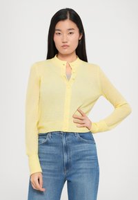 Lys gul cardigan med rund krage, knappet front, lange ermer og ribbestrikkede mansjetter, paret med blå høyt livjeans.