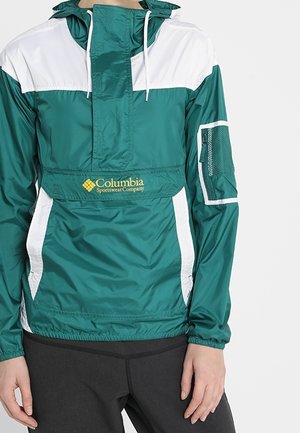 Zielona i biała wiatrówka Columbia z przednią kieszenią, siateczkową kieszenią na lewym ramieniu, noszona z czarnymi spodniami, ręce luźno opuszczone wzdłuż ciała.