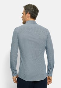 Mann mit kurzen, lockigen Haaren, der ein eng anliegendes, langärmliges, blau gemustertes Hemd trägt und mit dem Rücken zu einem schlichten weißen Hintergrund steht.