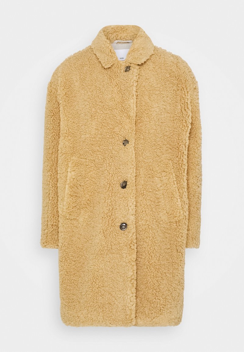 Manteau teddy beige avec manches longues, col, fermeture à boutons et deux poches latérales, en tissu doux et texturé.