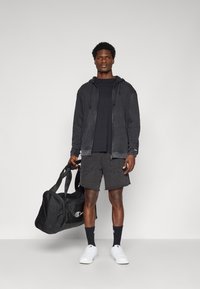 Schwarze Kapuzenjacke mit Kordelzügen, schwarzes T-Shirt, schwarze Shorts, weiße Sneakers, trägt eine schwarze Tasche. Merkmale sind eine lockere Passform und ein strukturiertes Material.
