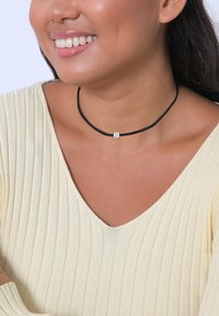Sort snor choker halskæde med en lille, klar, facetteret ædelsten som accent. Tekstureret, blødt materiale, båret over en lysfarvet top.