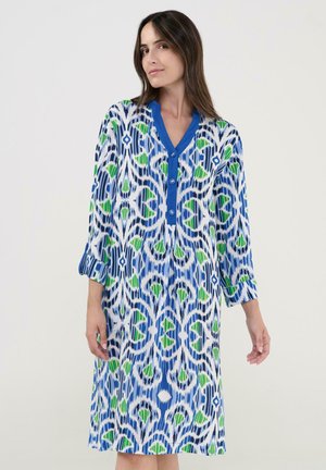 Mujer con vestido hasta la rodilla de patrón azul, verde y blanco, con mangas tres cuartos y escote con botones azules.