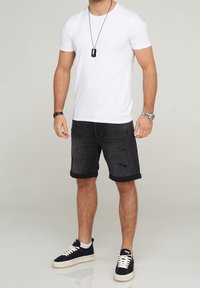 Camiseta blanca de manga corta, pantalones cortos de mezclilla oscura con puños enrollados y detalles desgastados, combinados con zapatillas negras y un collar de placa de perro.