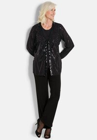 Schwarze Strickjacke mit Pailletten in einem geometrischen Muster; kombiniert mit einem schwarzen Top und einer Hose; hohe Schuhe rundet das Outfit ab.