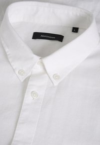 Chemise blanche à boutons avec un col pointu, trois boutons visibles et une étiquette intérieure noire portant la mention "Matinique", taille large.