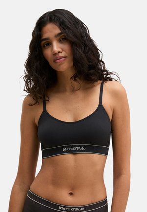 Woman with curly dark hair wearing a black ribbed bralette and matching underwear with "Marc O'Polo" logo on elastic bands.