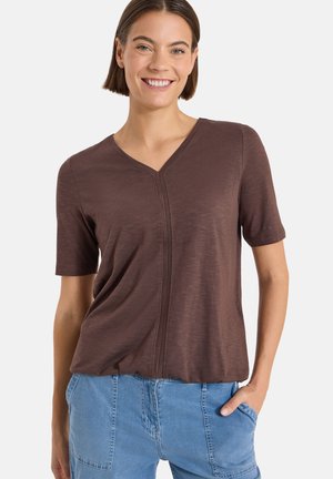 Femme souriante portant un haut marron à manches courtes et col en V ainsi qu’un jean bleu clair, avec la main droite dans la poche, sur fond blanc.