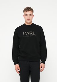 Mladý muž oblečený v čiernej mikine s logom "KARL LAGERFELD", stojaci na pozadí jednoduchého bieleho pozadia, čelí dopredu.