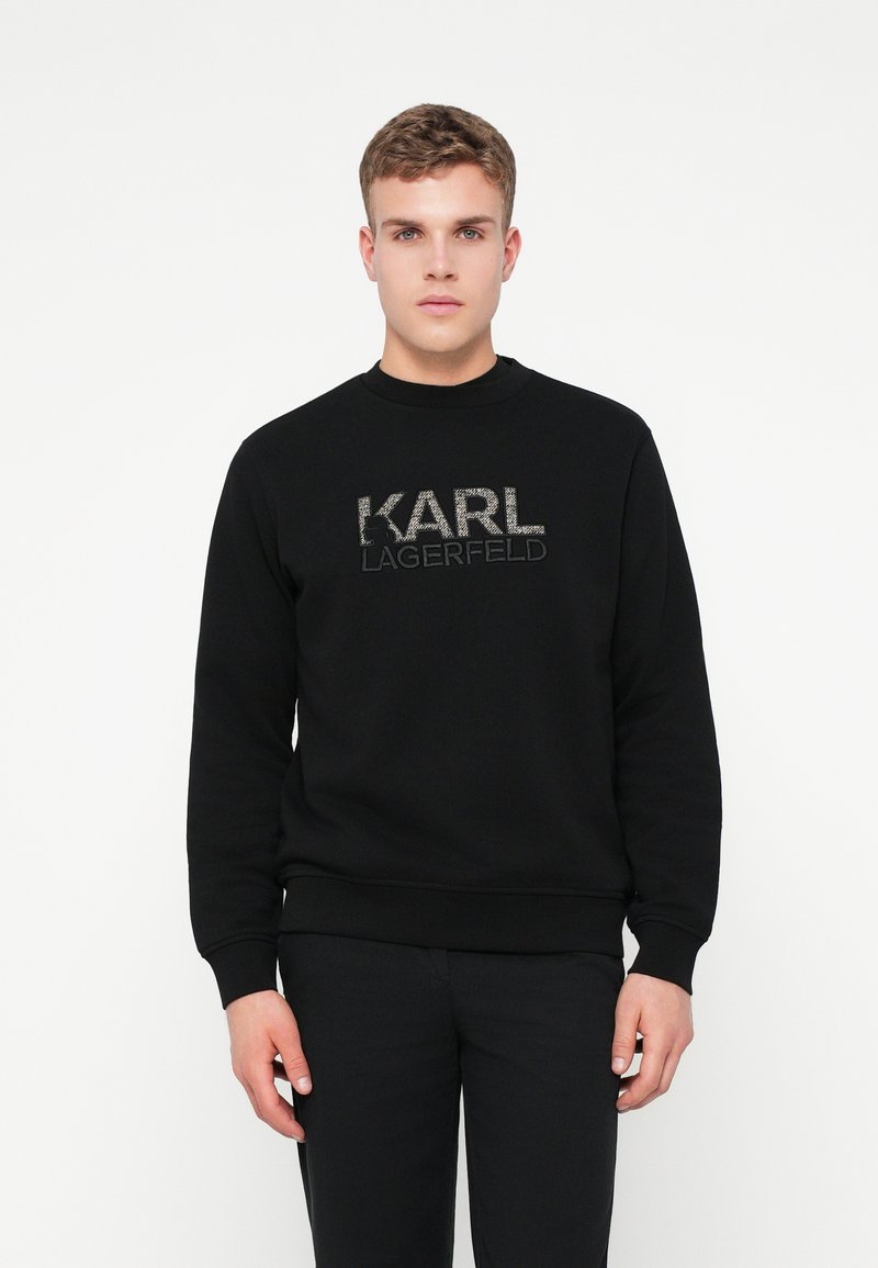 Mladý muž oblečený v čiernej mikine s logom "KARL LAGERFELD", stojaci na pozadí jednoduchého bieleho pozadia, čelí dopredu.