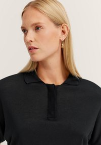 Schwarzes Strick-Polohemd mit strukturiertem Kragen und Knopfleiste. Das Model trägt goldene Ohrhängerringe und hat glattes, blondes Haar.